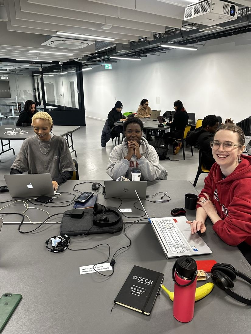 Three students smiling at a shared table with laptops during a class.