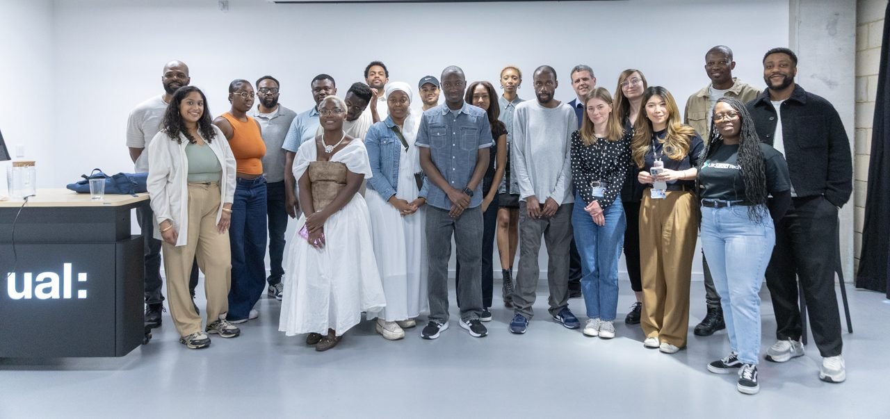 Group photo of around 25 Peckham DAZ students, tutors and partners standing together in a UAL teaching space, next to a podium with the UAL logo.