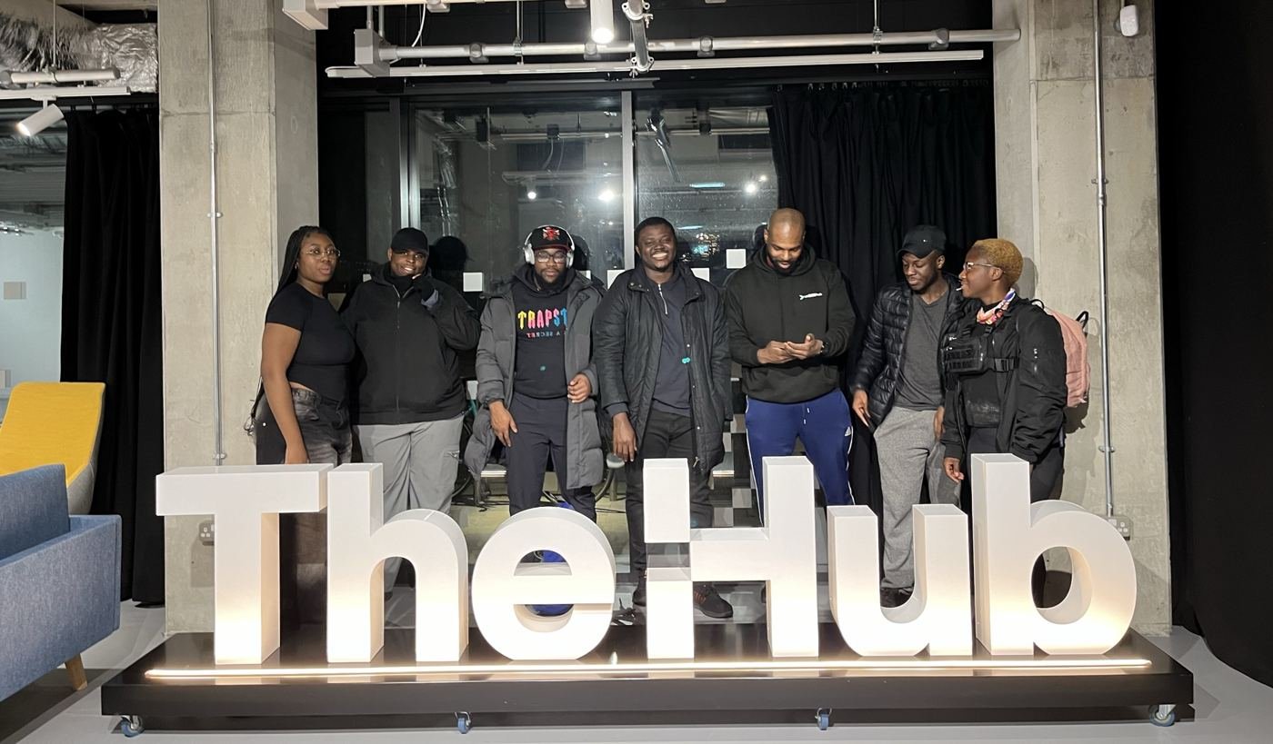 A group of Peckham DAZ students and coaches standing behind a large illuminated sign reading 'The Hub'.
