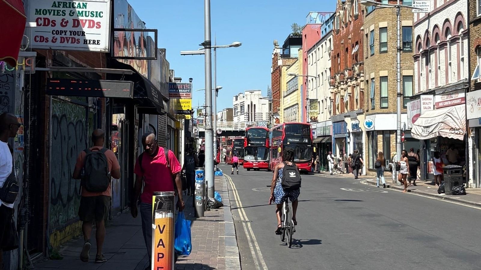 Rye Lane in Peckham on a sunny day: shoppers on the pavement, a cyclist, red double-decker buses and rows of independent shops.