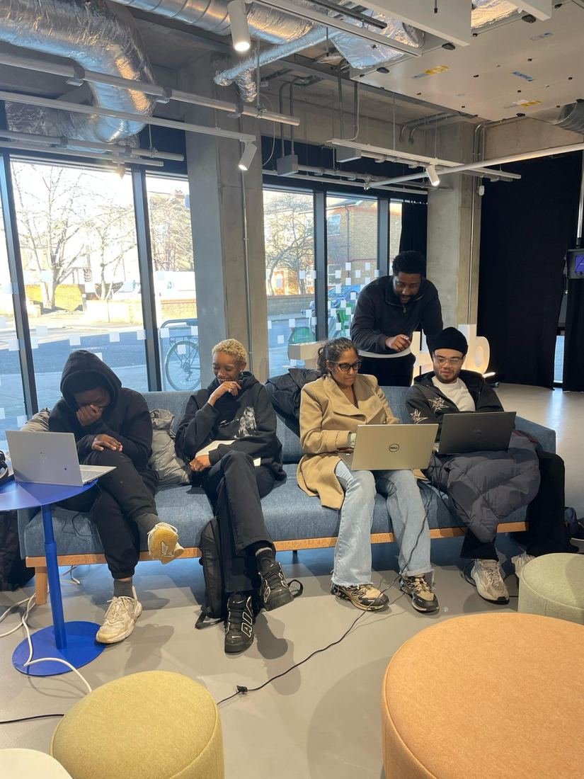A tutor leans in to help students working on laptops on a sofa.