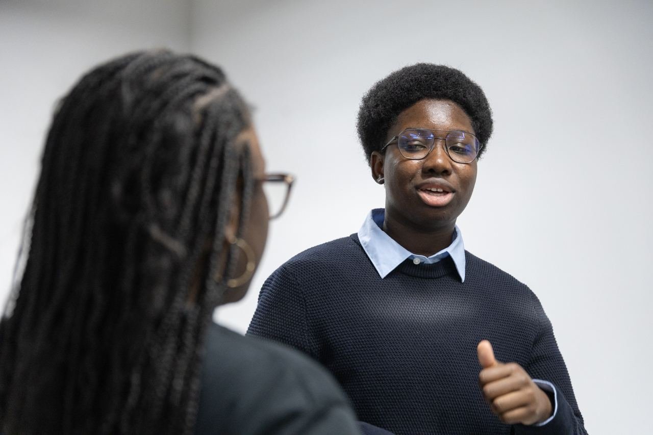 A student in glasses and a navy jumper makes a point during a discussion.
